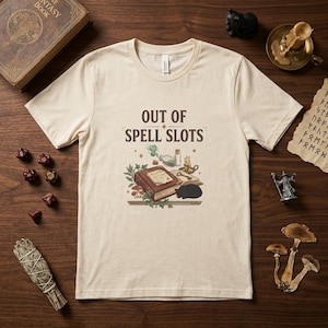 May include: Cream-colored t-shirt with the text "OUT OF SPELL SLOTS" above a graphic of a spell book, potion bottles, and a black cat. The design has a fantasy theme, with mushrooms and stars.