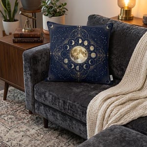 May include: A square decorative pillow with a navy blue background and a gold celestial design. The design features a full moon surrounded by phases of the moon and geometric shapes. The pillow is on a dark gray velvet couch.