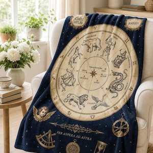May include: A navy blue throw blanket featuring a celestial map design with zodiac symbols and constellations. The blanket has a circular chart in the center with the text "CAELI ATLAS" and "PER ASPERA AD ASTRA". The blanket is draped over a white armchair.