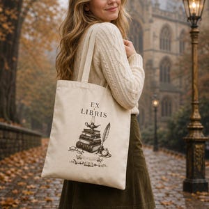 Op de afbeelding: Beige draagtas met de tekst "EX LIBRIS" en een afbeelding van boeken, een lamp en een veer. De tas wordt gedragen door een persoon die een crèmekleurige trui en een groene rok draagt. Op de achtergrond een bakstenen pad en een gebouw.