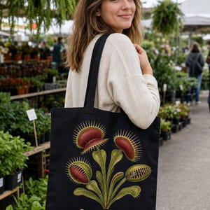 May include: A black tote bag featuring a botanical illustration of a Venus flytrap with red and green leaves. Below the plant, the text reads Dionaea muscipula and CARPE MUSCAM in white lettering, accompanied by two small golden bee illustrations.