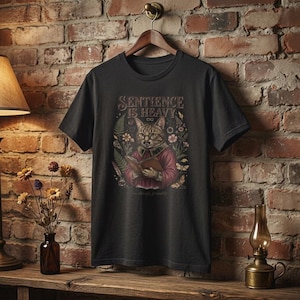 Może przedstawiać: Ciemnoszara koszulka z napisem "SENTIENCE IS HEAVY" nad ilustracją kota w okularach i czerwonej kurtce, trzymającego książkę. Projekt zawiera kwiaty i motyle. Koszulka wisi na drewnianym wieszaku.