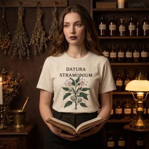 May include: A cream-coloured t-shirt with the words "Datura Stramonium" and a botanical illustration. The image shows a woman holding a book, with a background of apothecary bottles and dried herbs.