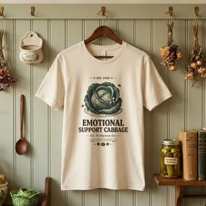 Könnte beinhalten: Cremefarbenes T-Shirt mit einer Grafik eines grünen Kohls und dem Text "EMOTIONAL SUPPORT CABBAGE". Das Shirt hängt an einem Holzbügel. Weitere Artikel im Bild sind ein Salzbehälter, Trockenblumen und ein Glas Gurken.