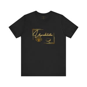 May include: Black t-shirt with a gold graphic design. The design includes the word "Unpredictable" in a stylized font, along with an eye and a crescent shape. The graphic is enclosed within a rectangular frame.