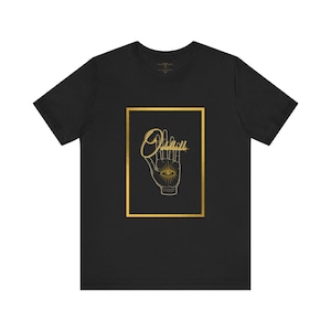 May include: Black t-shirt with a gold rectangular graphic. The graphic features a hand with an eye in the palm and the word "Oddball" in a script font. The design is centered on the shirt.