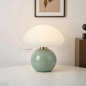 Può includere: Una moderna lampada da tavolo con base rotonda verde menta e paralume bianco a forma di fungo. La lampada ha un dettaglio color ottone e il testo "LUMIERE BY ORIYAN" è visibile. La lampada è su una superficie bianca.