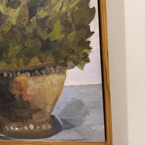 Könnte beinhalten: Ölgemälde eines Blumenarrangements in einer dekorativen Urne. Das Kunstwerk zeigt eine Vielzahl von grünem Laub und blauen Blumen. Die Urne hat eine warme braune Farbe und steht auf einer hellblauen Oberfläche. Das Gemälde ist in einem hellbraunen Holzrahmen gerahmt.