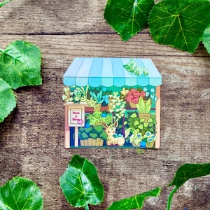 K&ouml;nnte beinhalten: Ein farbenfroher Aufkleber, der ein Blumengesch&auml;ft mit einer blauen Markise und verschiedenen Pflanzen und Blumen darstellt. Ein Schild lautet "Plant & Bloom". Ein Zeichentricktier ruht auf der Markise. Der Aufkleber befindet sich auf einer Holzoberfl&auml;che mit gr&uuml;nen Bl&auml;ttern.