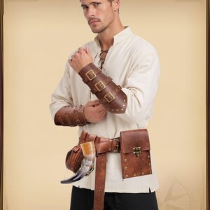 K&ouml;nnte beinhalten: Braune Leder-Armsch&uuml;tzer mit drei Schnallen und Nieten. Ein passender brauner Lederg&uuml;rtel mit einer Tasche und einem Horn sind ebenfalls sichtbar. Das Outfit erinnert an mittelalterliche oder Fantasy-Kleidung.