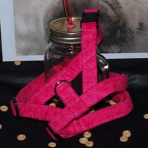 May include: A pink quilted dog harness with a black buckle and a silver ring. The harness is lying on a black surface with some cereal scattered around it.