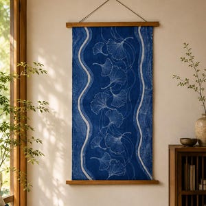 May include: A rectangular wall hanging featuring a deep blue fabric panel with white line art of ginkgo leaves and wavy vertical borders. The textile is secured between two horizontal wooden rods and suspended by a thin cord from the top.