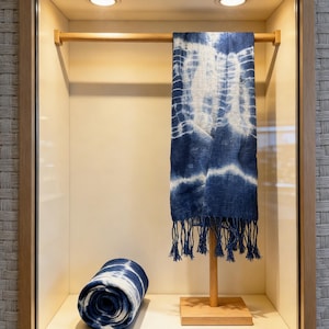May include: A blue and white tie-dye scarf with fringe edges hangs on a wooden display stand, while a matching rolled-up scarf sits on the base. The fabric features a distinct indigo-colored pattern with white organic shapes and lines.