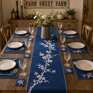 May include: A dark blue table runner and matching placemats featuring a white cherry blossom branch pattern. The set includes square coasters with the same floral design, arranged on a wooden dining table with white plates and clear glasses.
