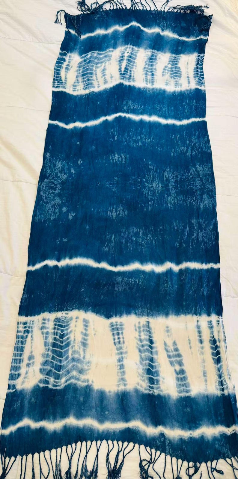 May include: A rectangular scarf featuring a tie-dye pattern in deep blue and white. The design consists of horizontal bands of solid blue alternating with white sections that display intricate, repeating chevron-like textures. The ends have fringe.