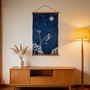 May include: A navy blue wall hanging with a white floral design and a full moon. The artwork is displayed above a wooden cabinet with a white vase and dried plants. A wooden floor lamp with a yellow lampshade is to the right.
