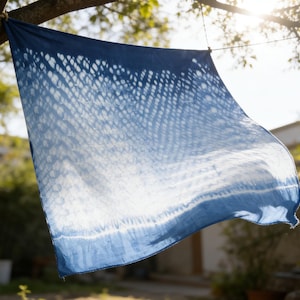 May include: A square, indigo-dyed textile, hanging outdoors. The fabric features a gradient effect, transitioning from a deep blue at the top to white at the bottom. The design includes a dotted pattern. The textile is suspended from a tree branch.