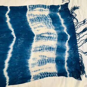 May include: A rectangular scarf featuring a tie-dye pattern with alternating bands of deep indigo blue and white. The fabric displays a textured, crinkled appearance with vertical stripes and a fringe of loose blue threads along the short edge.