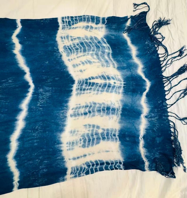 May include: A rectangular scarf featuring a tie-dye pattern with alternating bands of deep indigo blue and white. The fabric displays a textured, crinkled appearance with vertical stripes and a fringe of loose blue threads along the short edge.