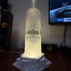 May include: A lamp made from a Belvedere vodka bottle. The bottle is clear with a frosted tree and building design. The base is a white, geometric design. The bottle is lit from within, creating a warm glow.