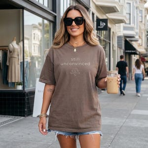 Op de afbeelding: Een bruin t-shirt met de woorden "still unconvinced" in het wit gedrukt. Het shirt wordt gedragen met een spijkerbroek en een zonnebril. De persoon houdt een glas ijskoffie met een rietje vast.