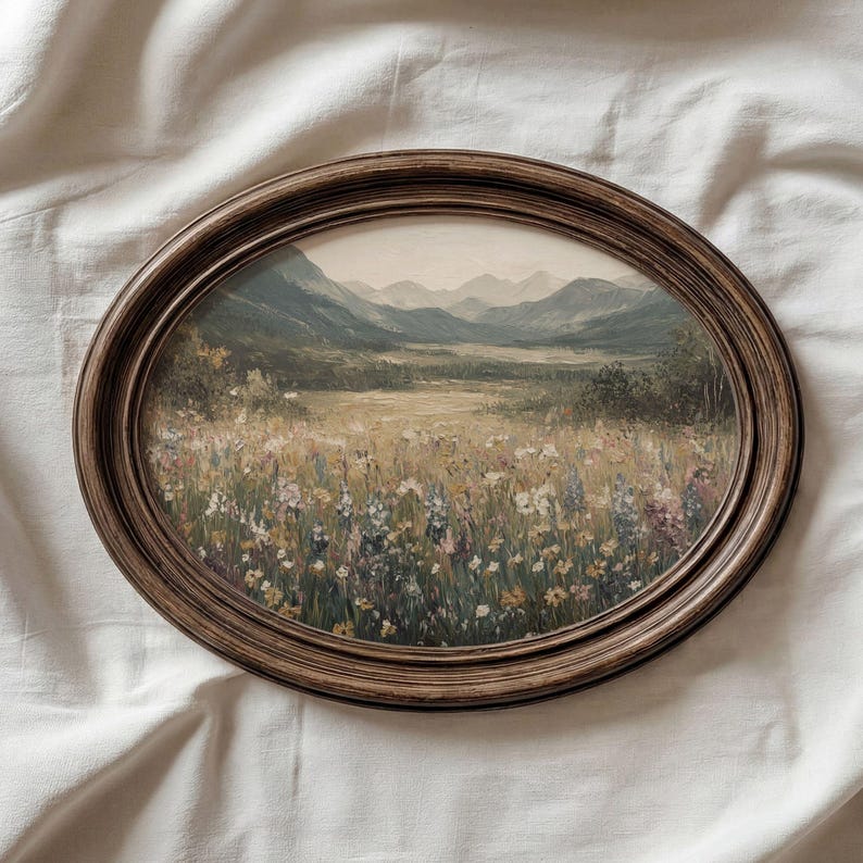 May include: An oval framed landscape painting. The artwork shows a field of wildflowers, with hills and mountains in the background. The frame is dark brown wood. The painting evokes a vintage aesthetic, suitable for home decor.