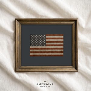 May include: Framed print of the American flag with a distressed, vintage appearance. The flag features red and white stripes and a field of white stars on a dark blue background. The frame is a dark, ornate gold color. The text "EMERSONS FINE ART" is at the bottom.