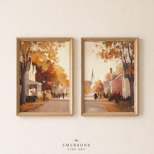 May include: Two framed vertical art prints depicting autumn street scenes with houses, trees with orange and brown foliage, and figures walking along a path. The left print shows a house with a porch, while the right print features a church steeple.