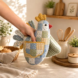 May include: A quilted oven mitt shaped like a chicken, featuring a patchwork design of blue, green, and yellow floral and gingham fabrics. The mitt has a mustard yellow comb, a red beak, a black eye, and a small pocket on the side of the body.