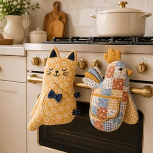 May include: Two fabric oven mitts hanging on a stove handle. One is a yellow cat with a daisy pattern, a small blue bow, and dark blue ears. The other is a chicken shape with a patchwork design of blue, yellow, and orange floral and gingham squares.