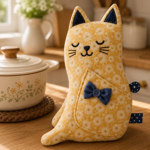May include: A cat-shaped fabric oven mitt featuring a yellow floral pattern, dark blue inner ears, and a small blue bow with a button on the front pocket. The design includes embroidered closed eyes and whiskers, with a blue polka dot tail detail.