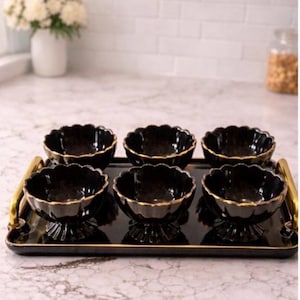 Puede incluir: Un juego de seis cuencos de cerámica negra con bordes festoneados y ribete dorado, dispuestos sobre una bandeja negra a juego con asas doradas. Los cuencos se exhiben sobre una encimera de mármol.