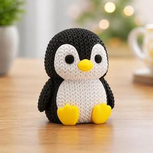 Puede incluir: Una pequeña figura de pingüino con una superficie texturizada en blanco y negro que imita el tejido. El pingüino tiene dos ojos negros redondos, un pico amarillo brillante y dos patas palmeadas amarillas en la base. Cuerpo y cabeza redondos.