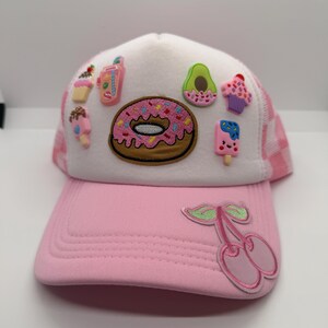 May include: Pink and white trucker hat with a pink peak and a white mesh back. The hat is decorated with a donut, avocado, cupcake, and popsicle patches. A cherry patch is on the front.