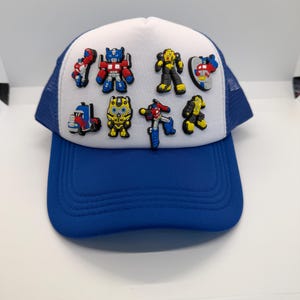 May include: A blue and white trucker hat with several colourful character embellishments. The hat has a blue peak and mesh back, with a white front panel decorated with various robot figures. The characters are in red, blue, yellow, and black.
