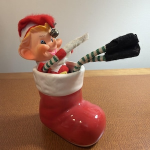 Peut inclure: Une figurine de chaussette de Noël en céramique rouge vif avec un revers blanc, contenant une petite poupée elfe. L'elfe porte un bonnet rouge avec une clochette argentée, une tenue rouge et blanche, et de longues jambes fines rayées vert et blanc.