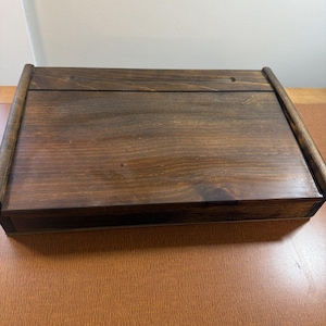 May include: A rectangular wooden box with a dark brown stained finish and a smooth, flat lid. The box features visible wood grain patterns and rounded edges along the sides, presenting a simple, solid construction with a horizontal seam.