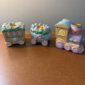 May include: Three pastel-colored Easter train candle holders. The train cars are decorated with flowers, eggs, and bows. The candle holders are made of ceramic and are designed to hold small candles. The train is on a brown surface.