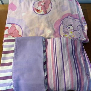 Puede incluir: Un juego de ropa de cama con temática de Ositos Cariñositos, con una combinación de colores rosa y morado. La sábana superior tiene gráficos de Ositos Cariñositos y formas de corazón. Las fundas de almohada son de color lavanda sólido y a rayas en tonos de morado, rosa y blanco.