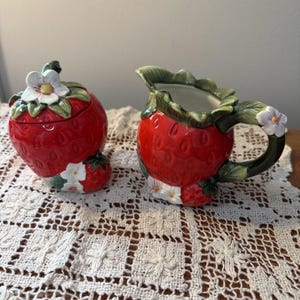 May include: A ceramic strawberry-shaped sugar bowl with a lid and a matching creamer. The sugar bowl has a white flower on top and a small strawberry at the base. The creamer has a handle and a white flower on the handle.