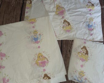Puede incluir: Un juego de ropa de cama blanca con un patrón repetitivo de princesas Disney, incluyendo a Bella, Cenicienta y Aurora. Cada personaje está enmarcado dentro de una corona floral circular de rosas rosas y hojas verdes sobre la tela.
