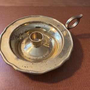 May include: A vintage brass candle holder with a circular base and a central cup for the candle. The holder has a handle and a decorative, scalloped edge. The brass has a warm, aged patina, suggesting it is an antique.