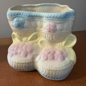 May include: A vintage ceramic planter shaped like two baby boots. The planter is painted in pastel shades, with blue and yellow accents, and pink floral details. The boots are joined by yellow ribbons.