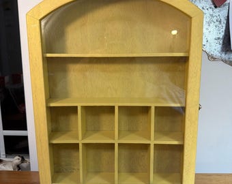 May include: A yellow wooden display case with an arched top and a clear glass front door. The interior features one full-width shelf at the top and eight smaller square compartments arranged in two rows of four at the bottom for organizing items.
