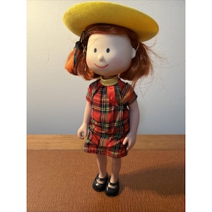 May include: A vintage doll with red hair, wearing a red plaid dress, black shoes, and a yellow hat. The doll has a painted face with a small smile and is standing on a wooden surface. The doll is a collectible item.