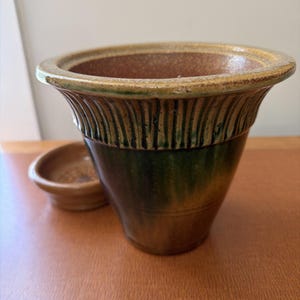 May include: A ceramic plant pot with a flared rim and vertical ribbed detailing. The exterior features a gradient glaze transitioning from a golden-brown top to a deep forest green base. A matching small, circular, unglazed terracotta saucer sits nearby.