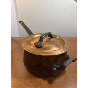 以下が含まれることがあります： 蓋付きの丸い銅製の鍋で、暗い色の金属製ハンドルが付いています。鍋の片側には長いハンドルが伸びており、反対側には短いハンドルが付いています。蓋には中央にハンドルがあり、酸化や摩耗の跡が見られます。