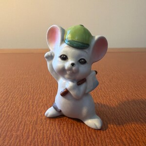 May include: A ceramic mouse figurine featuring a light gray body with pink-tinted ears, wearing a green baseball cap. The mouse is posed standing upright, holding a small brown stick-like object in its hands, with a cheerful facial expression.