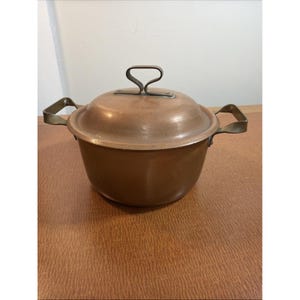 May include: A round copper pot with a matching lid and two brass-toned metal handles on the sides. The lid features a central looped handle. The pot has a smooth, metallic finish with visible signs of use and natural oxidation on the surface.