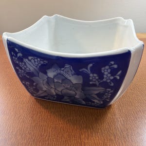 Puede incluir: Un cuenco cuadrado de cerámica con interior blanco y exterior azul profundo que presenta un patrón floral blanco. El cuenco tiene cuatro esquinas curvas y elevadas y una parte superior abierta, mostrando un acabado liso y brillante.
